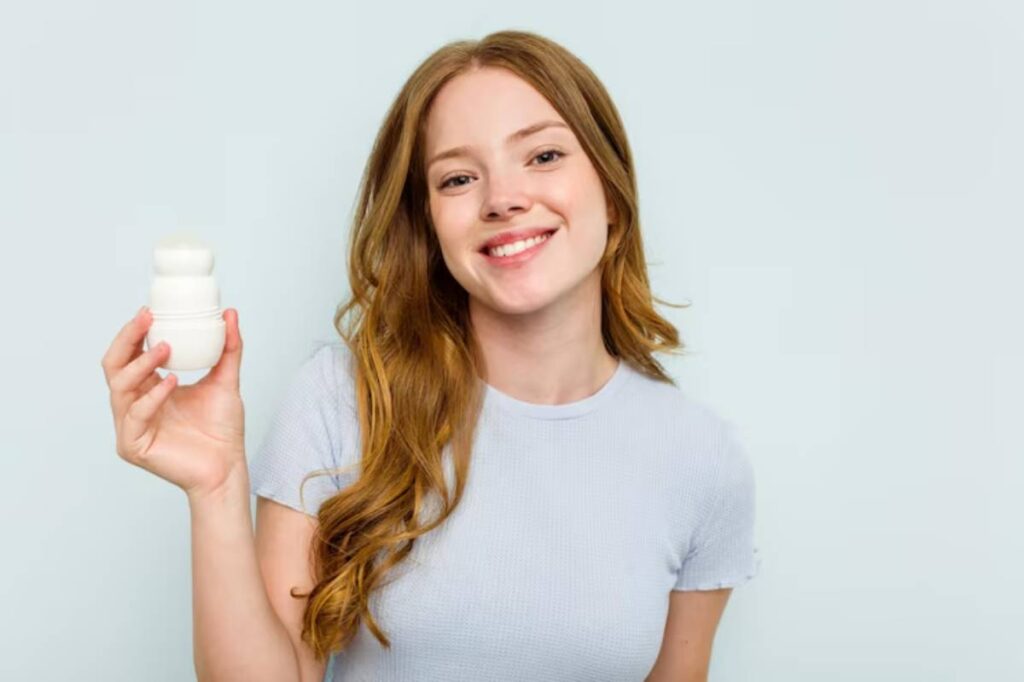 A woman with wavy, light brown hair holds probiotic, wearing a casual light gray shirt against a soft, pale background.