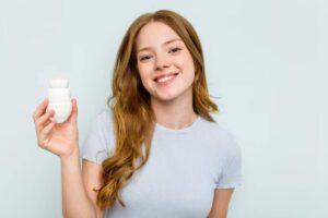A woman with wavy, light brown hair holds probiotic, wearing a casual light gray shirt against a soft, pale background.
