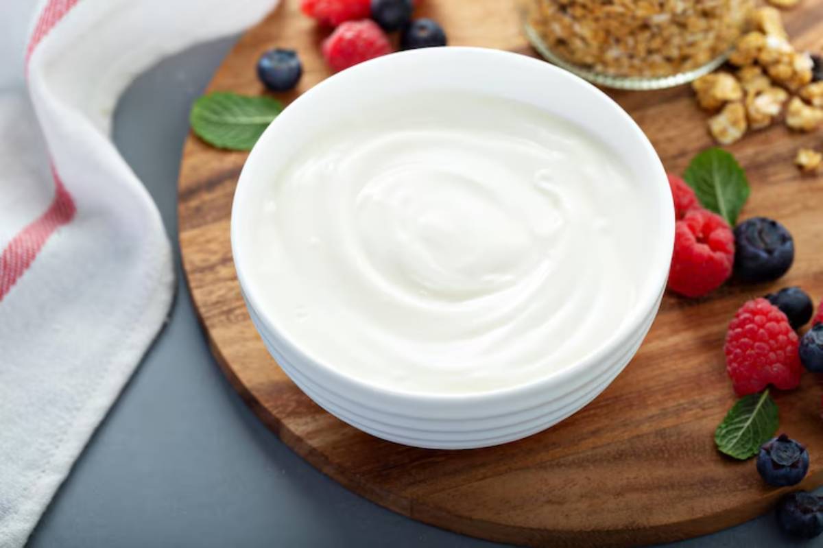 A creamy bowl of yogurt surrounded by fresh raspberries, blueberries, and mint on a wooden cutting board.
