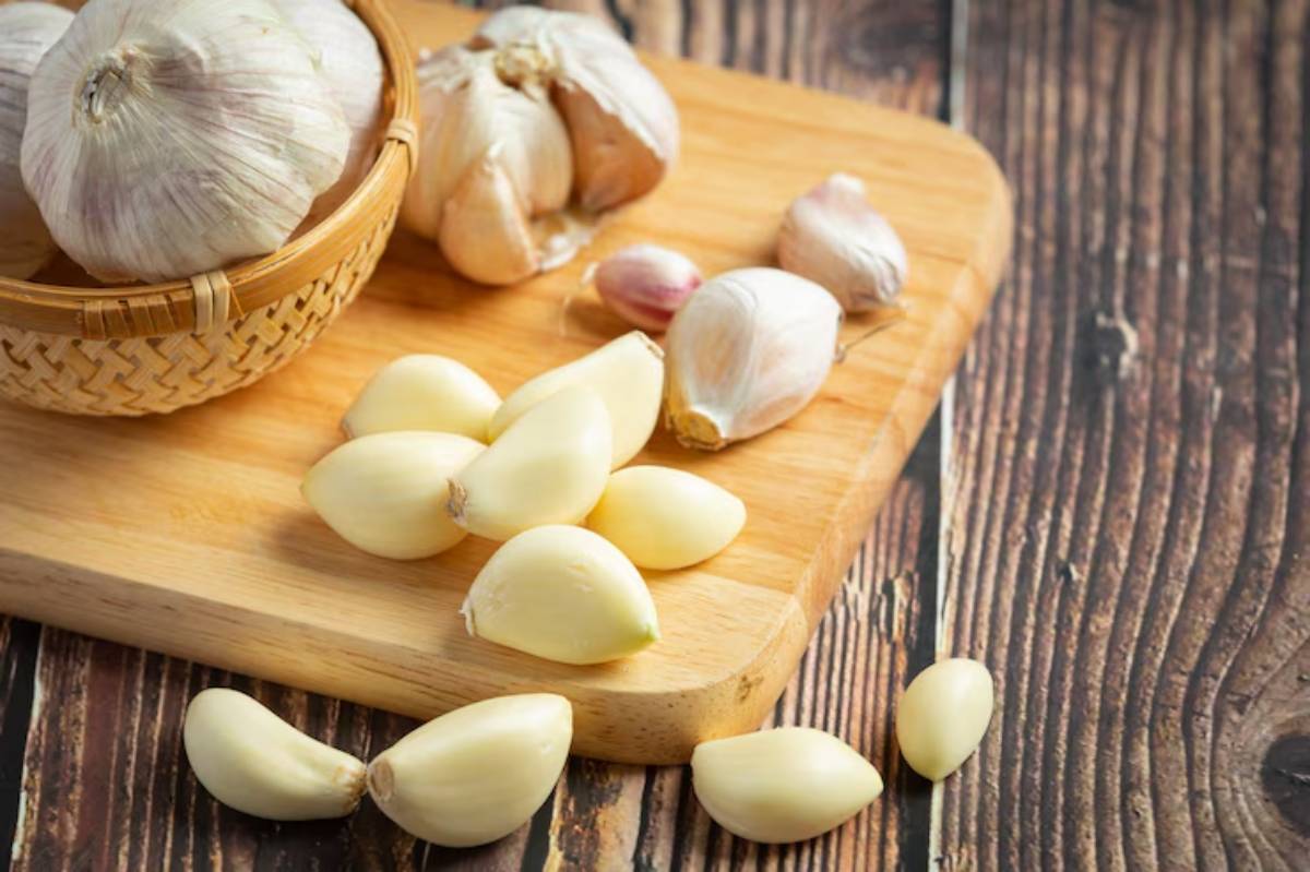 A wooden cutting board with several garlic cloves and a woven basket of whole garlic bulbs, on a rustic wooden surface.