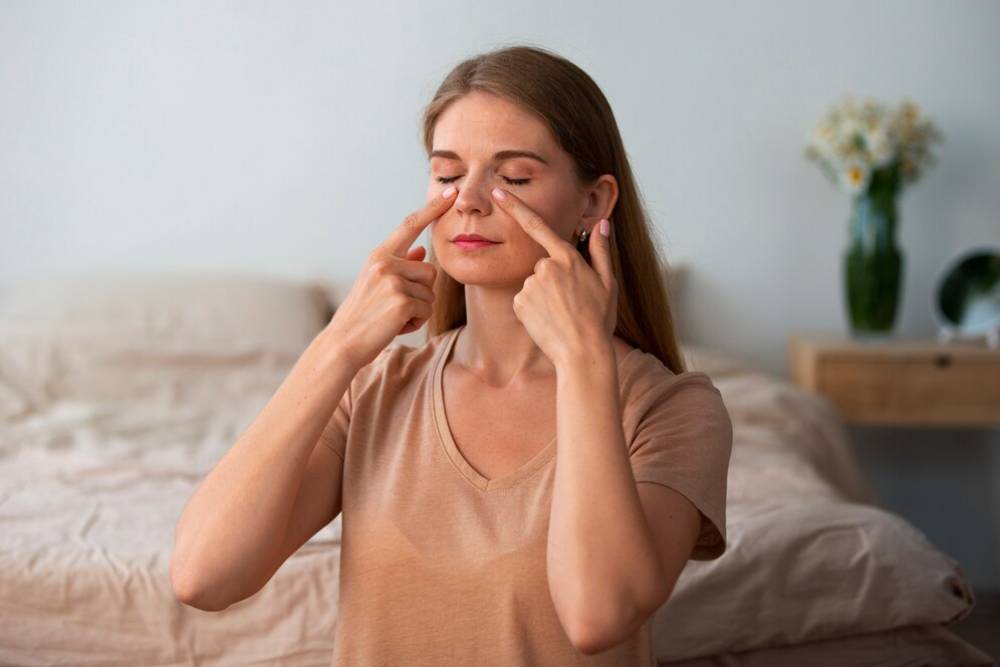 A woman in a beige t-shirt sits on a bed, doing facial yoga