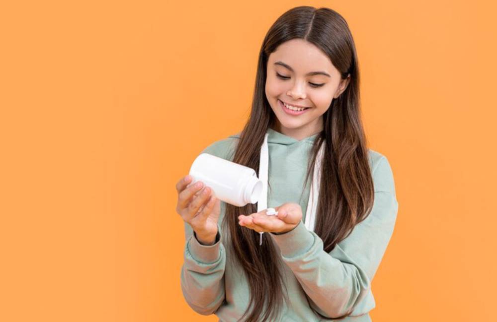 A person in a green hoodie holds a white pill bottle, pouring out two capsules into their palm, against a bright orange background.