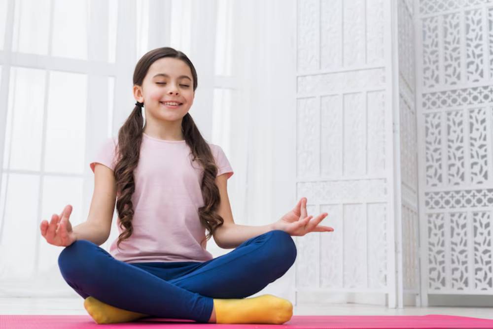 A child sits cross-legged on a pink mat, practicing meditation in a bright, serene indoor setting with decorative screens in the background.
