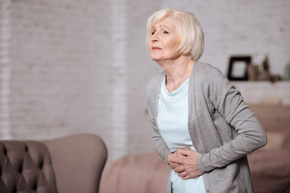 An elderly woman in a gray cardigan and light blue top clasps her stomach, appearing uncomfortable in a cozy living room setting.