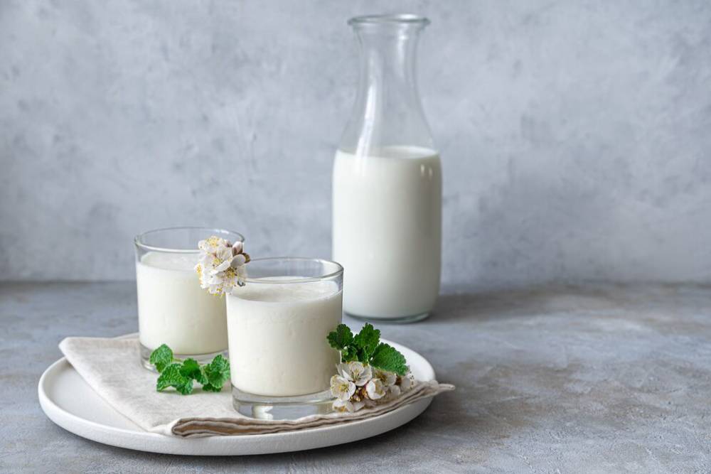 Two glasses of milk adorned with flowers and mint, accompanied by a milk bottle on a neutral grey background.