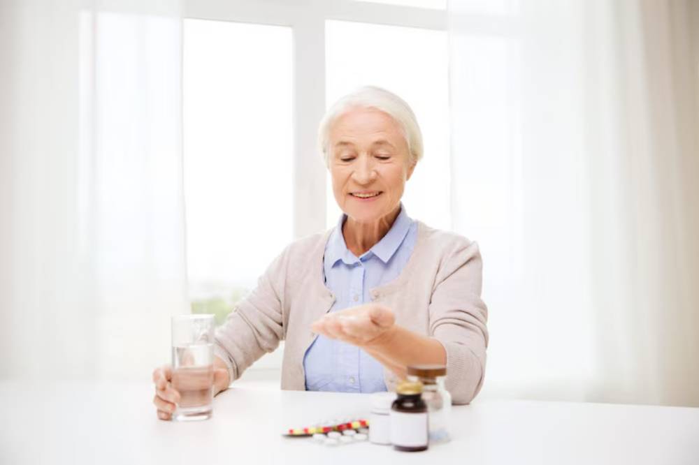 An elderly woman sits at a table with a glass of water, holding a handful of pills, surrounded by various medication containers.