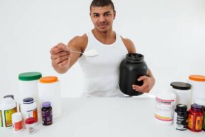 A person in a white tank top holds a black probiotics container and a spoon, surrounded by various dietary supplements on a table.
