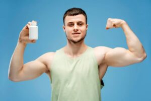 A muscular man in a light green tank top raises his arm to flex while holding a white supplement bottle against a blue background.