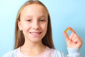 A child holds a capsule, standing against a soft blue background, wearing a light pink shirt and a cardigan.