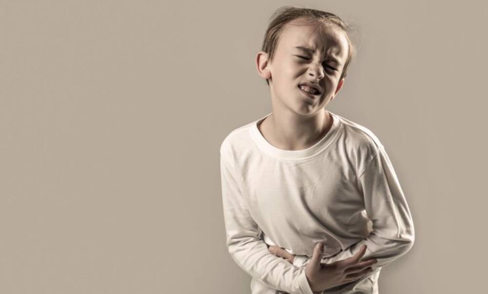 A child in a plain white shirt stands with hands on their stomach, showing signs of discomfort against a beige background.