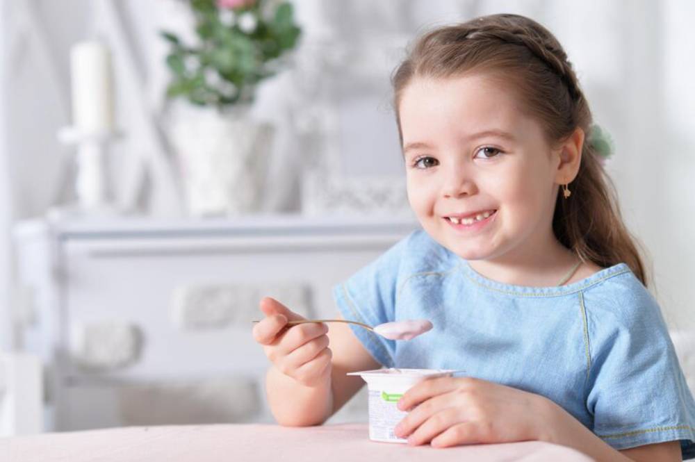 A child in a light blue shirt holds a spoon and yogurt container, enjoying a playful snack in a softly lit, cozy setting.