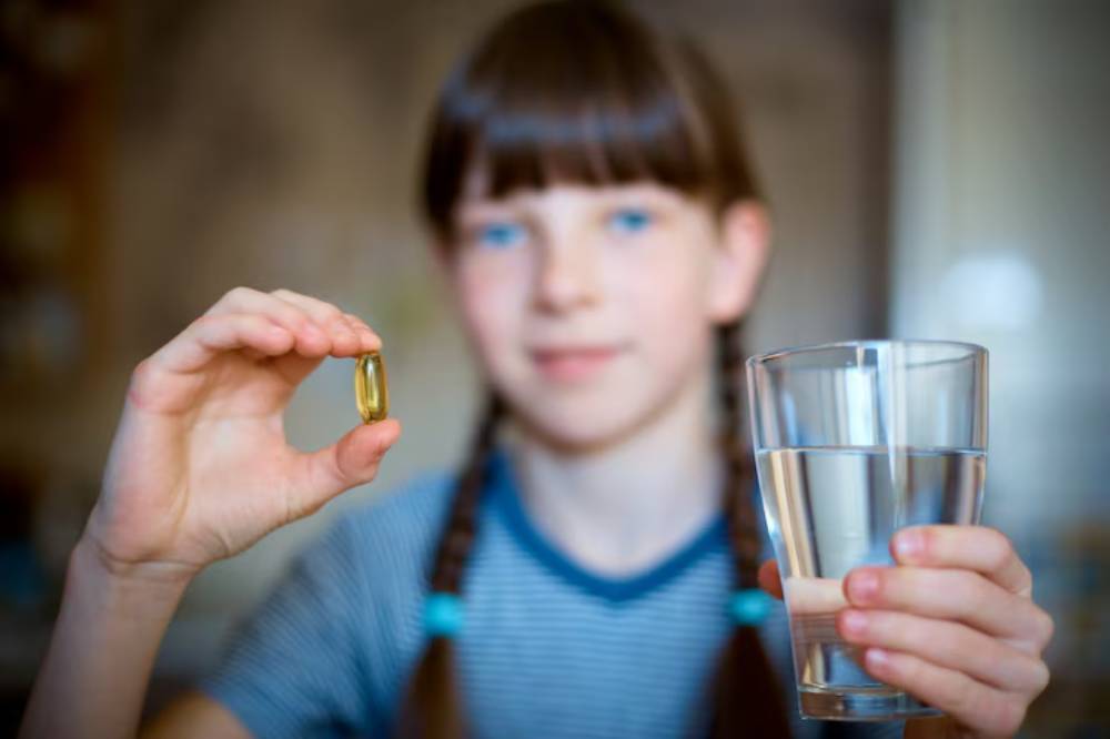 A child holds a golden supplement capsule in one hand and a glass of water in the other, preparing to take the supplement.