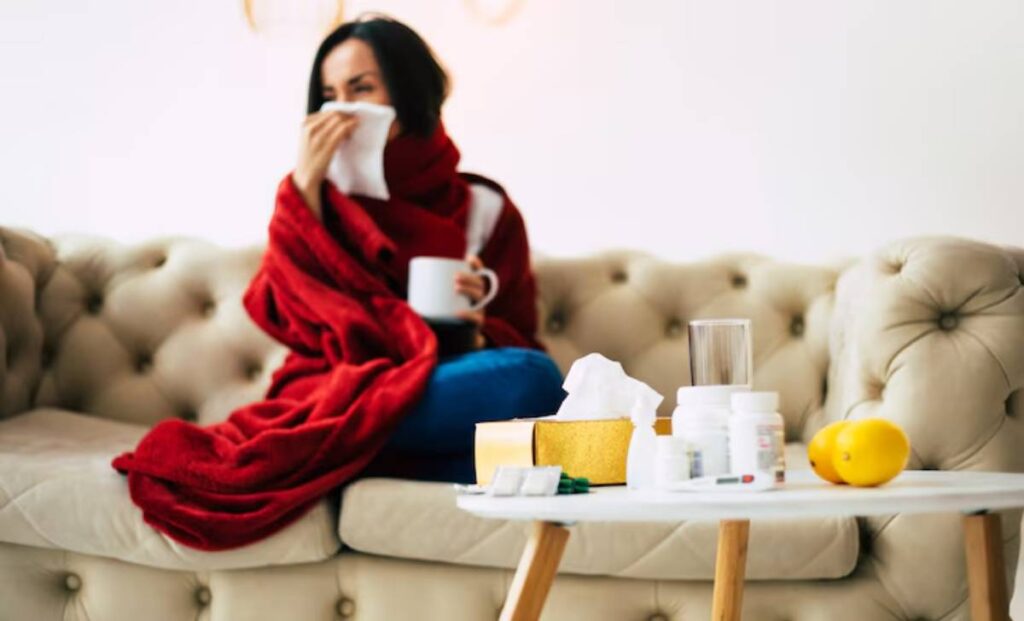 A person wrapped in a red blanket sits on a couch, holding a cup, with medicine and tissues on a nearby table.
