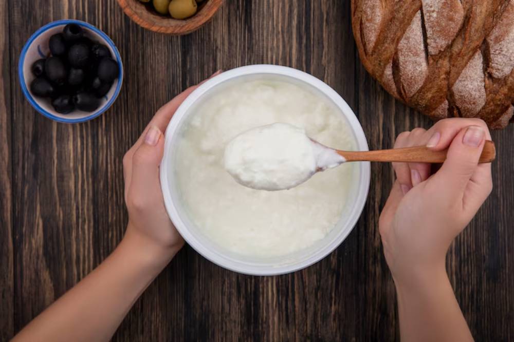 A person holding a wooden spoon scoops creamy yogurt from a white container, surrounded by olives and crusty bread on a wooden table.