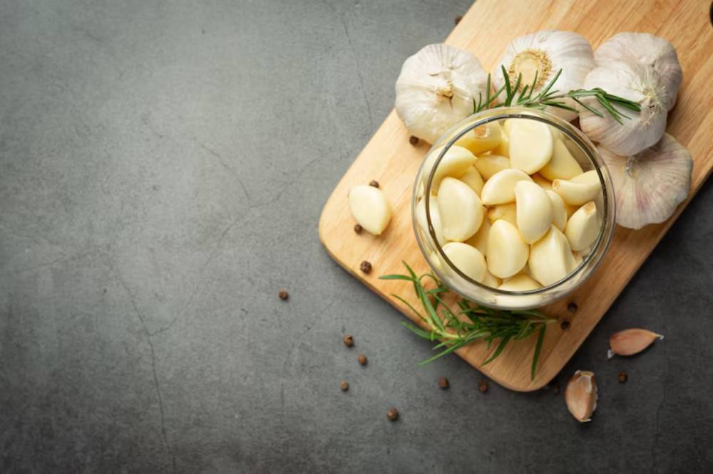 A glass jar filled with peeled garlic cloves sits on a wooden cutting board, surrounded by whole garlic bulbs and herbs.