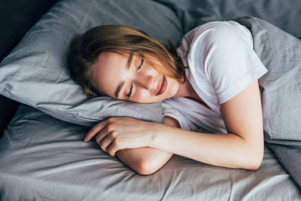A person sleeping on a gray bed, resting their head on a pillow, with arms wrapped around themselves in a serene atmosphere.