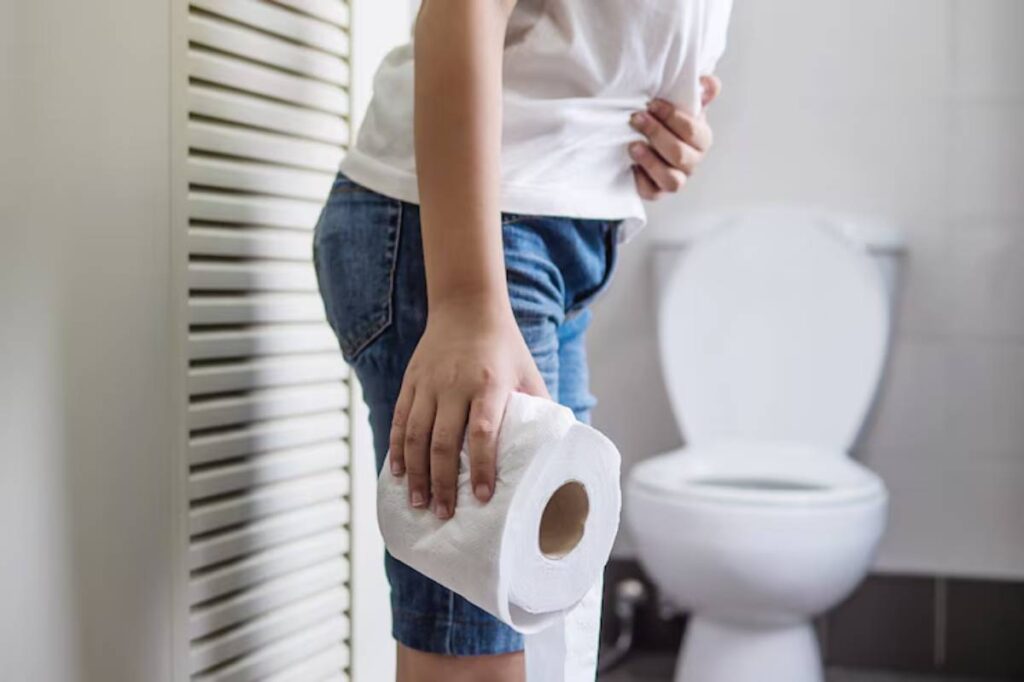 A person in a white shirt and denim shorts holds a roll of toilet paper, standing near a toilet in a bathroom setting.