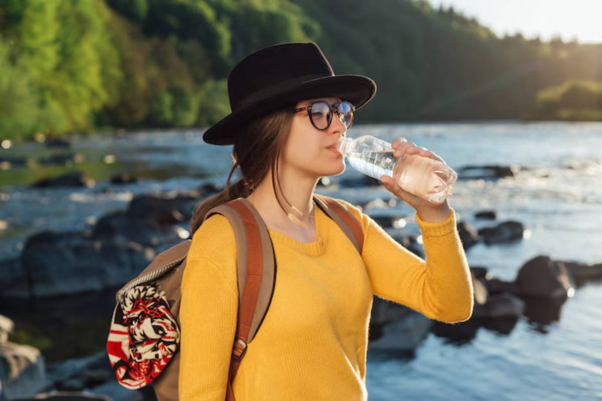 A person in a yellow sweater and hat drinks water by a tranquil river, surrounded by lush greenery and smooth rocks.