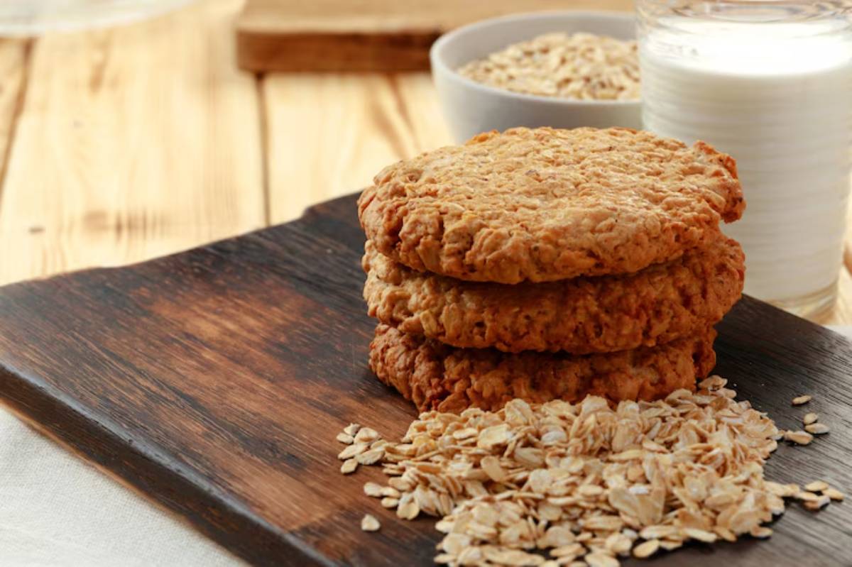 Three golden brown oat cookies stacked on a wooden board, surrounded by oats, with a glass of milk in the background.