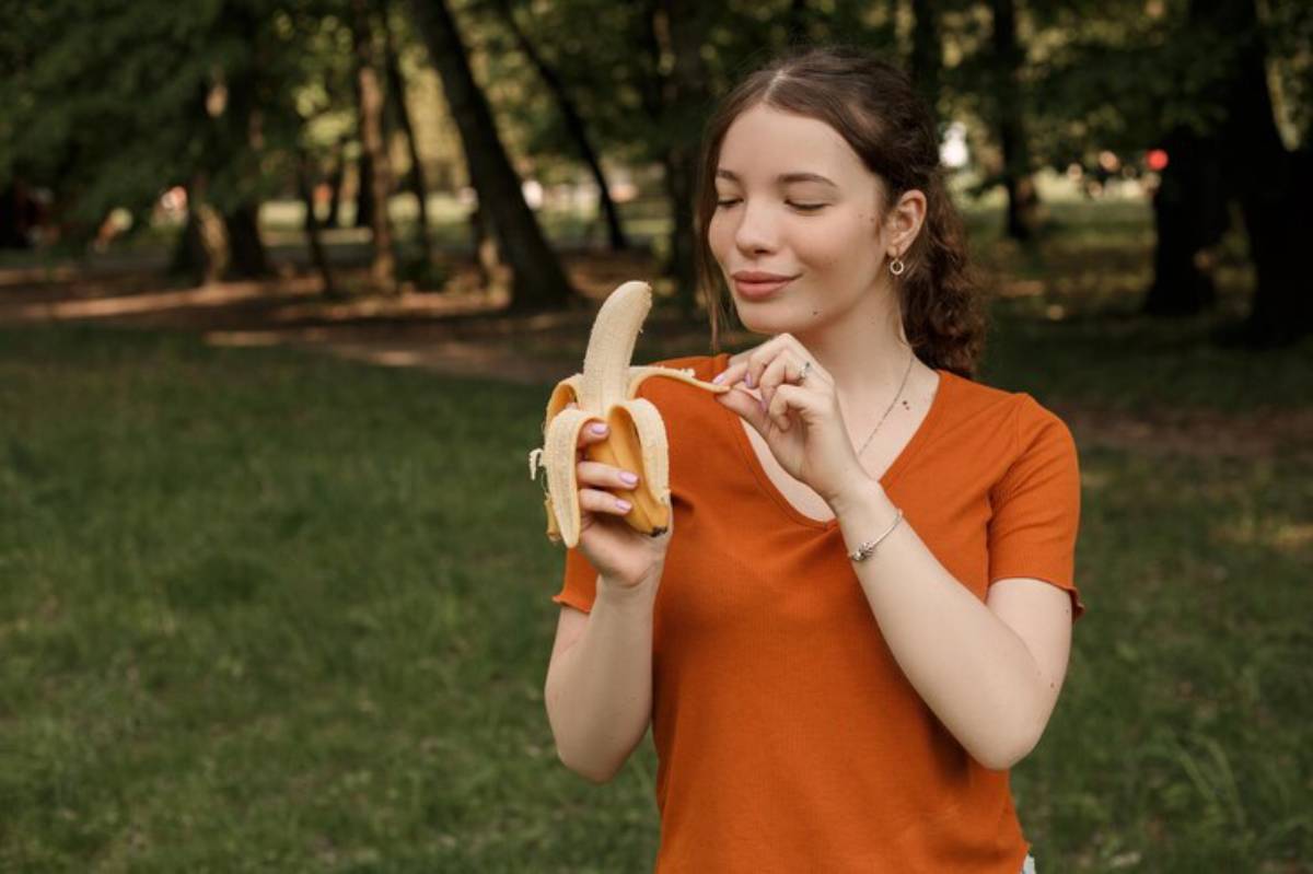 A person holding a peeled banana, standing in a green park surrounded by trees, wearing an orange shirt.