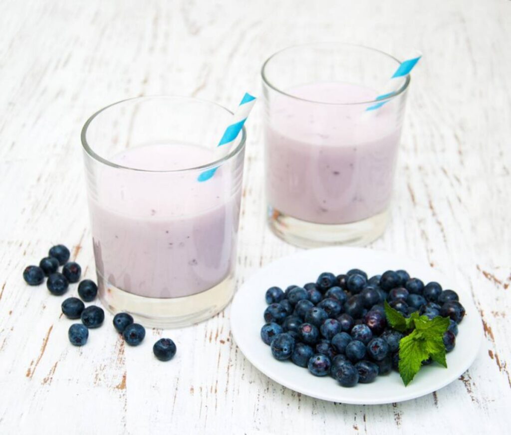 Two glasses of creamy pink blueberry smoothies with blue-striped straws, accompanied by a plate of fresh blueberries and mint.