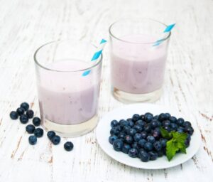 Two glasses of creamy pink blueberry smoothies with blue-striped straws, accompanied by a plate of fresh blueberries and mint.