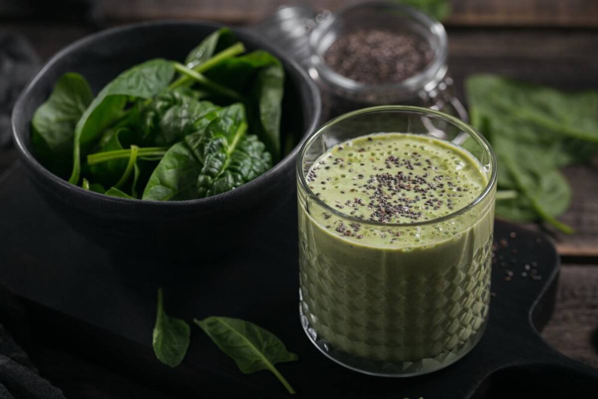 A glass of green smoothie topped with chia seeds sits beside a bowl of fresh spinach leaves on a dark wooden surface.