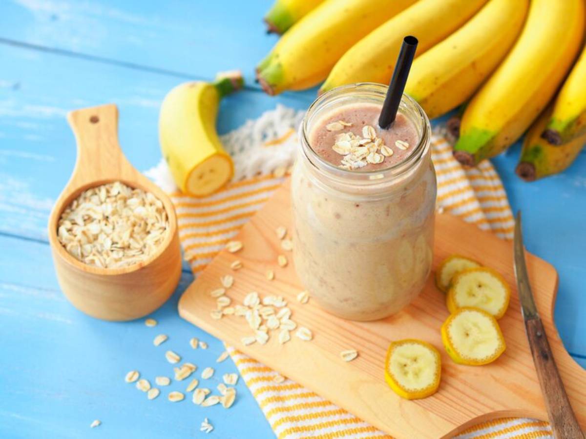 A jar of banana smoothie with oats, surrounded by sliced bananas and a scoop of oats on a wooden board against a blue background.