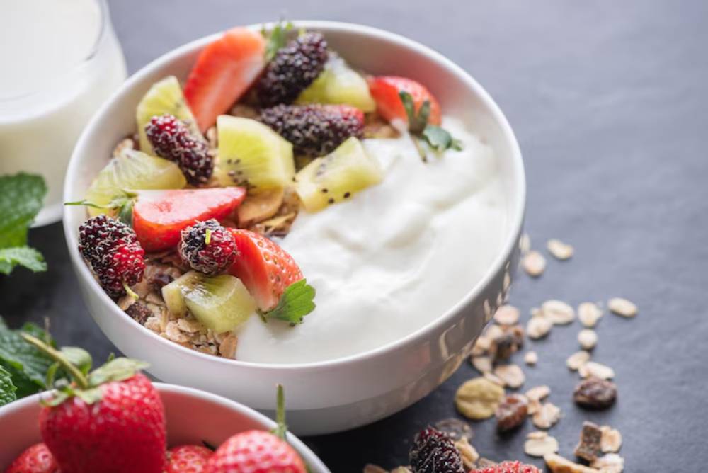 A bowl of yogurt topped with strawberries, kiwi, blackberries, and granola, with a glass of milk and fresh strawberries nearby.