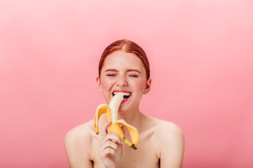 A person holds a peeled banana near their face against a bright pink background, showcasing a playful and artistic vibe.