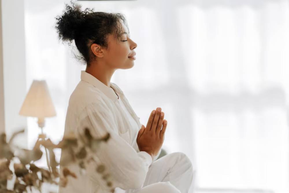 A person in white attire sits in a meditative pose with hands pressed together, surrounded by soft natural light and a cozy ambiance.