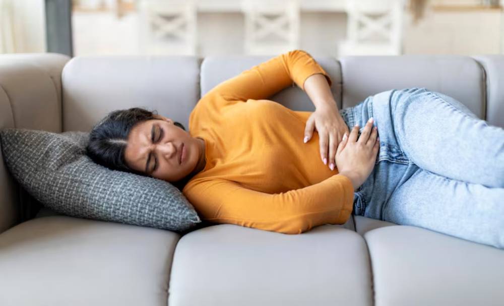 A person in an orange long-sleeve shirt lies  on a beige couch, resting with hands on their stomach.