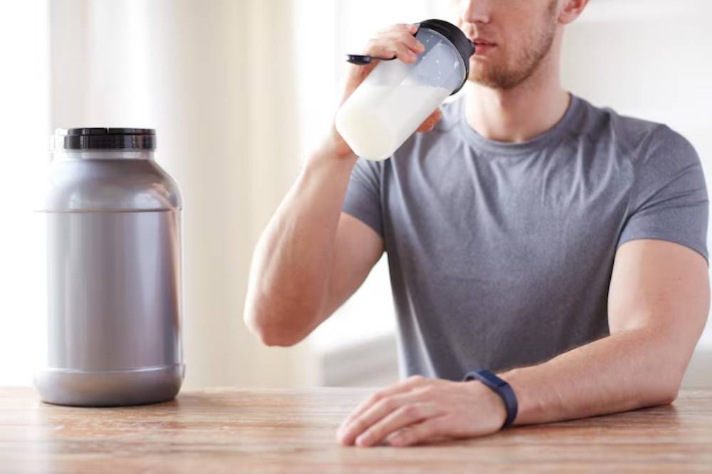 A person in a gray shirt is drinking from a bottle, with a large container of supplement.