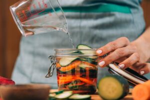 A hand pours water into a jar filled with colorful sliced cucumbers and peppers, with fresh vegetables surrounding the jar on a wooden surface.