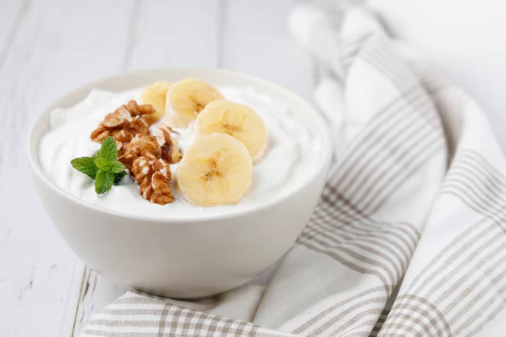 Bowl of creamy yogurt topped with banana slices, walnuts, and a fresh mint leaf, on a light checkered cloth background.