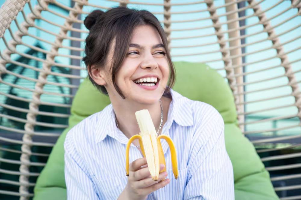 A person in a striped shirt holding a peeled banana, seated on a green cushion in a cozy, modern setting.