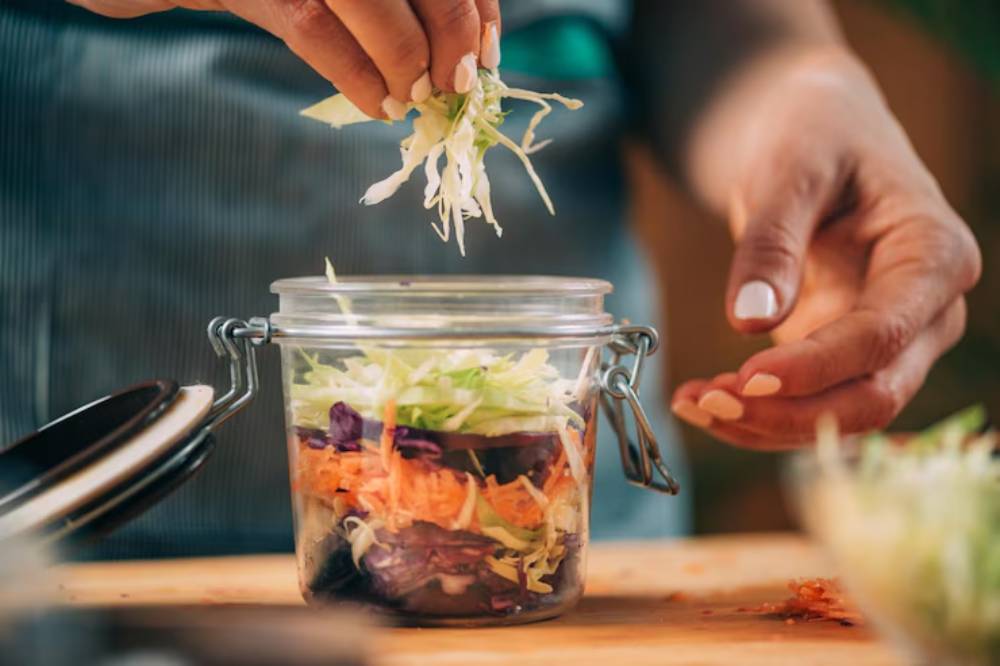 A hand adds shredded cabbage to a glass jar filled with colorful layers of vegetables salad ingredients on a wooden countertop.