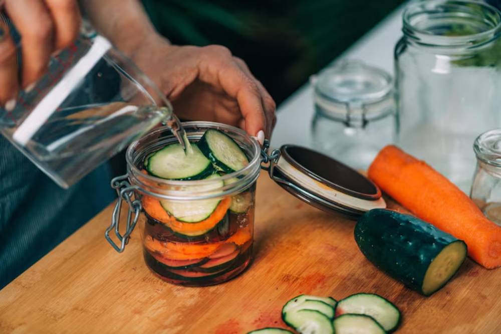 A person pours water into a jar filled with cucumber and carrot slices, with fresh vegetables on a wooden cutting board.