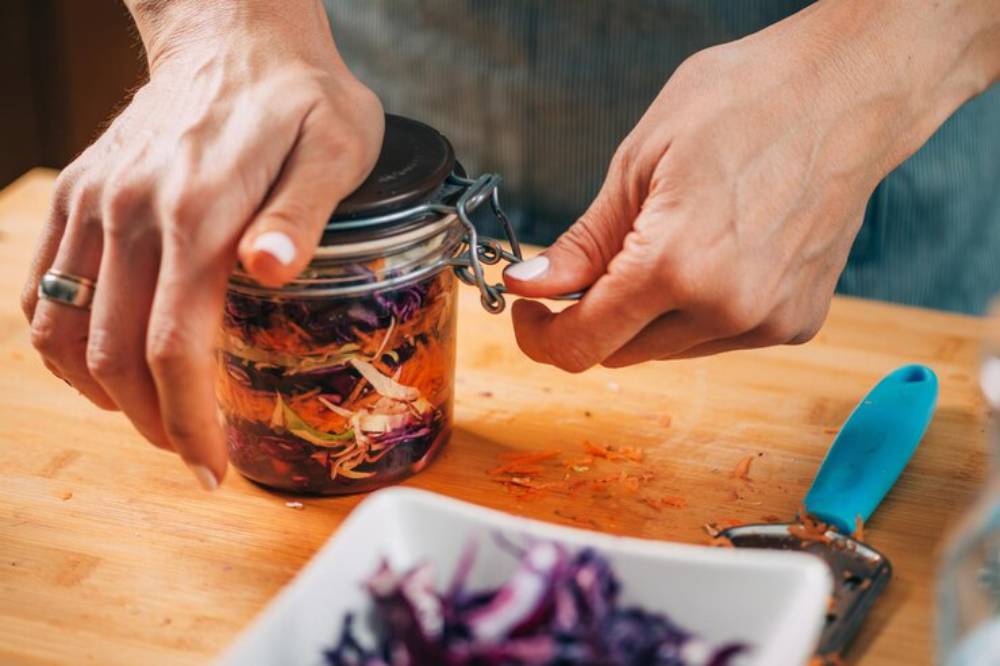 A person sealing a glass jar filled with colorful shredded vegetables, with a peeler and a bowl of veggies on a wooden countertop.
