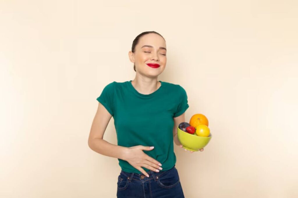 A person wearing a green shirt stands against a beige background, holding a bowl of colorful fruits while touching their stomach.