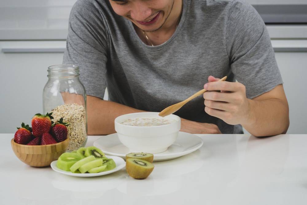 A man in a gray shirt enjoys a bowl of oatmeal, surrounded by fresh strawberries, kiwi, and oats on a bright kitchen table.