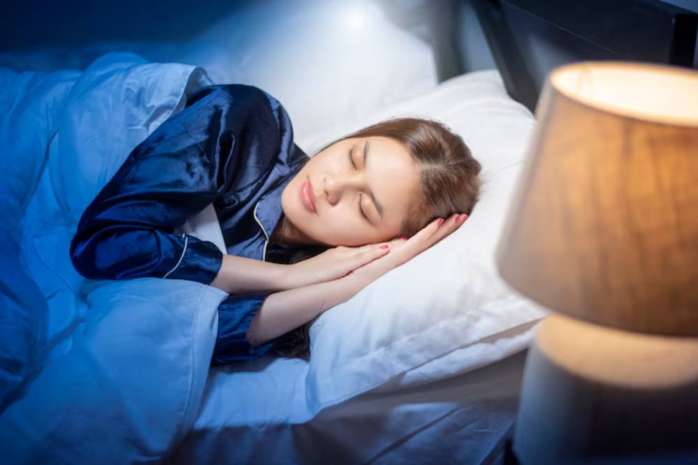 A person in a dark bedroom, covered with a white comforter, lying on a pillow beside a softly glowing lamp.