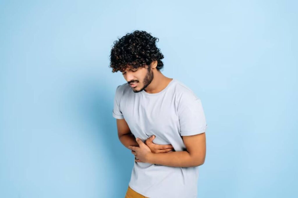 A person in a light gray t-shirt stands against a blue background, holding their stomach with a pained expression.