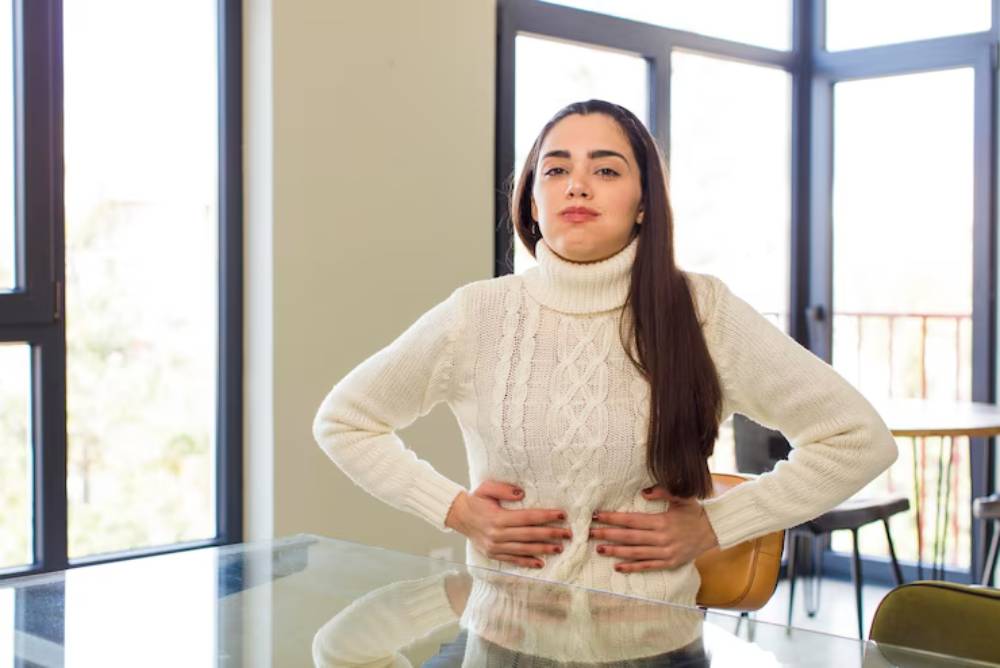 A person in a cozy white sweater stands at a table, with hands resting on their stomach, in a bright, modern interior.