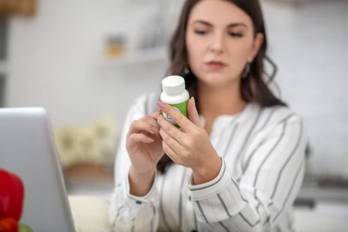 A person in a striped shirt holds a green and white supplement bottle, sitting at a desk with a laptop and vegetables nearby.