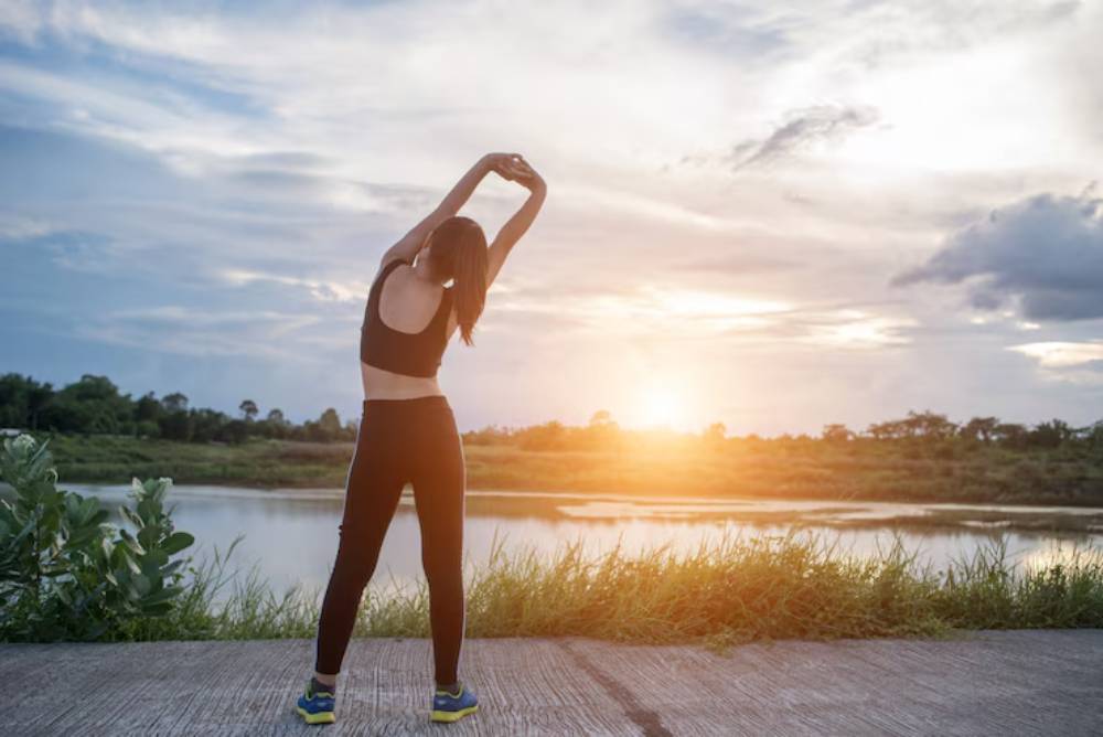 A woman with long hair stretches by a tranquil river at sunset, silhouette against a colorful sky, promoting fitness and tranquility.