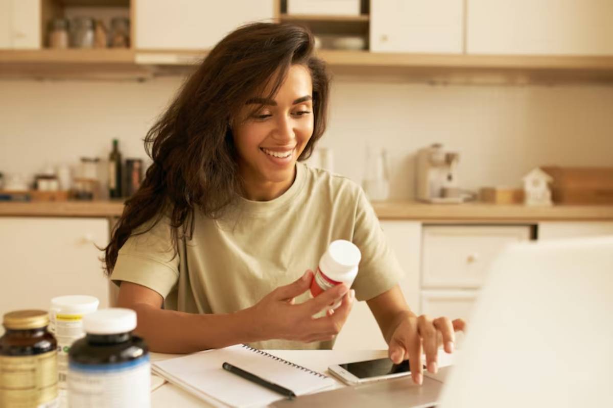 A person holding a red and white pill bottle, surrounded by various supplement bottles, while using a laptop on a kitchen table.