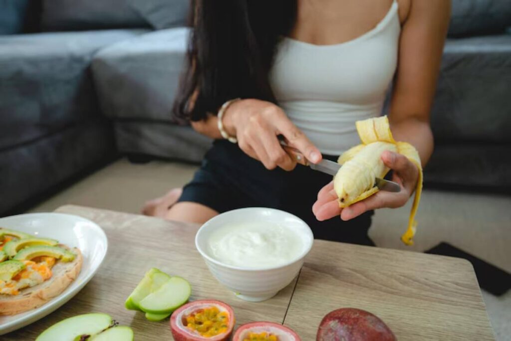 A woman peels a banana with a knife, sitting at a table with fresh fruits and a bowl of yogurt beside her.