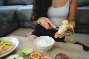 A woman peels a banana with a knife, sitting at a table with fresh fruits and a bowl of yogurt beside her.