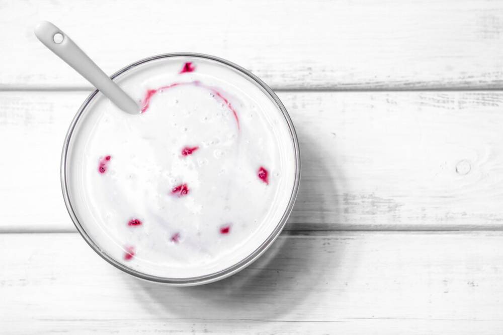 A bowl of creamy yogurt with small red raspberry pieces, topped with a silver spoon, on a white wooden surface.